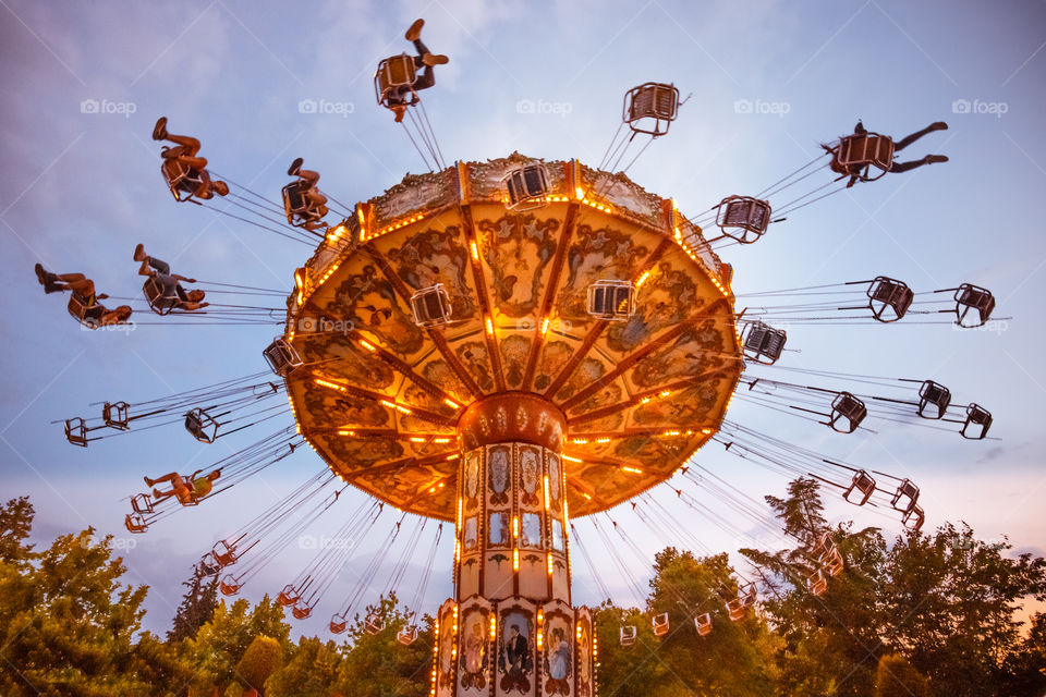 People have fun at the colorful chain flying swing carousel attraction in motion, at amusement park at night on blue sky background.