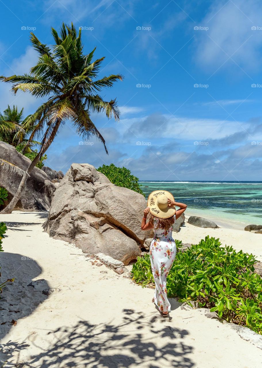 Young woman wearing sun hat and dress walking on exotic trpical sandy beach with palm trees and turquoise ocean