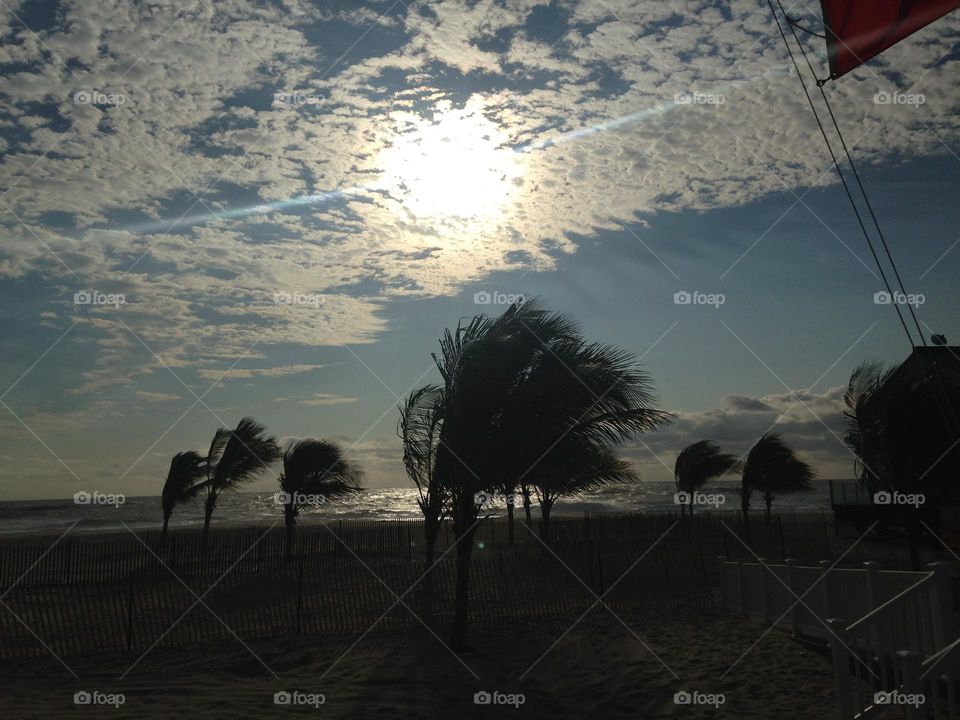 A stormy scene on a local beach. This photo was taken in the early morning in Point Pleasant Beach, NJ, but the beach is dark, except for a glow of a determined sun behind the clouds. The wind blows the trees and they tilt sideways.