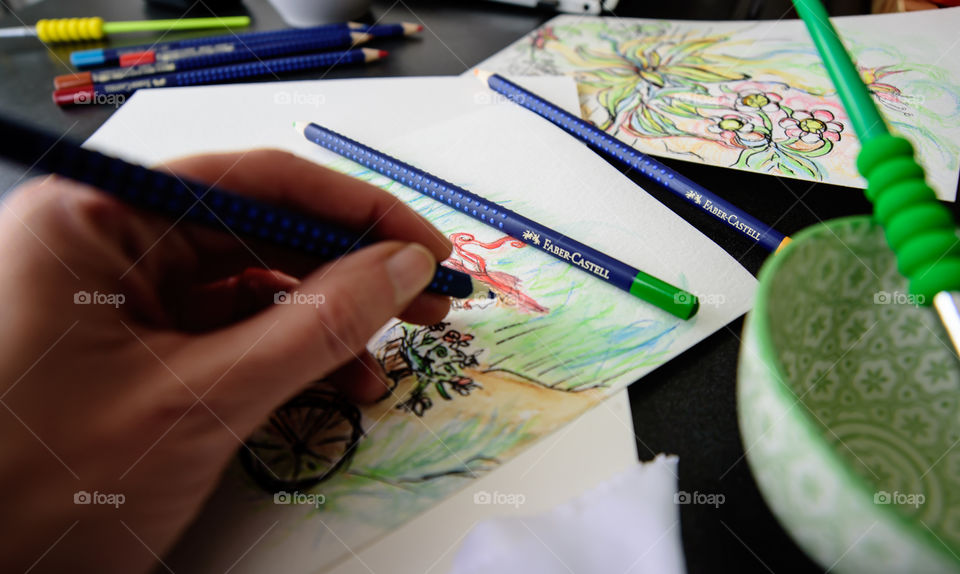 Closeup of hand holding Faber-Castell Aquarelle Colored watercolor pencils sketching girl on bicycle in countryside with sketchbook on table with laptop and coffee conceptual work life balance, creativity, hobby artist lifestyle photography