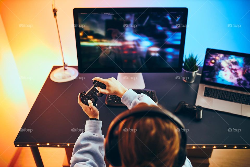 Teenager girl playing video game at home. Gamer holding gamepads sitting at front of screen. Streamer playing online in dark room lit by neon lights. Competition and having fun