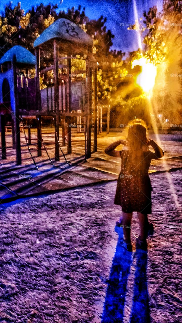 Light from a lamppost shines behind a playground in Athens at dusk and the colors dance mixing cold and warm colors
