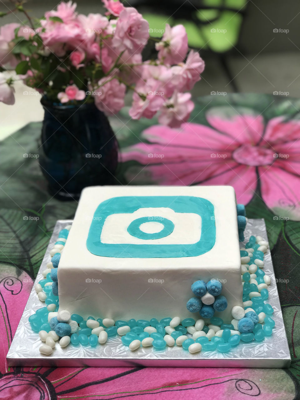A foap wedding cake with the logo made of white & turquoise fondant. Daisies of turquoise gummies & marshmallows on the side of the cake & turquoise & white jelly beans on the cake base. All on a green & pink table cloth with a vase of pink roses.