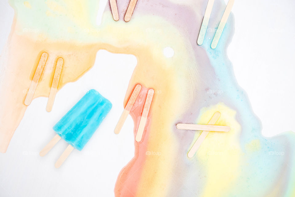 Flat lay of melted frozen popsicles forming a rainbow on a white background