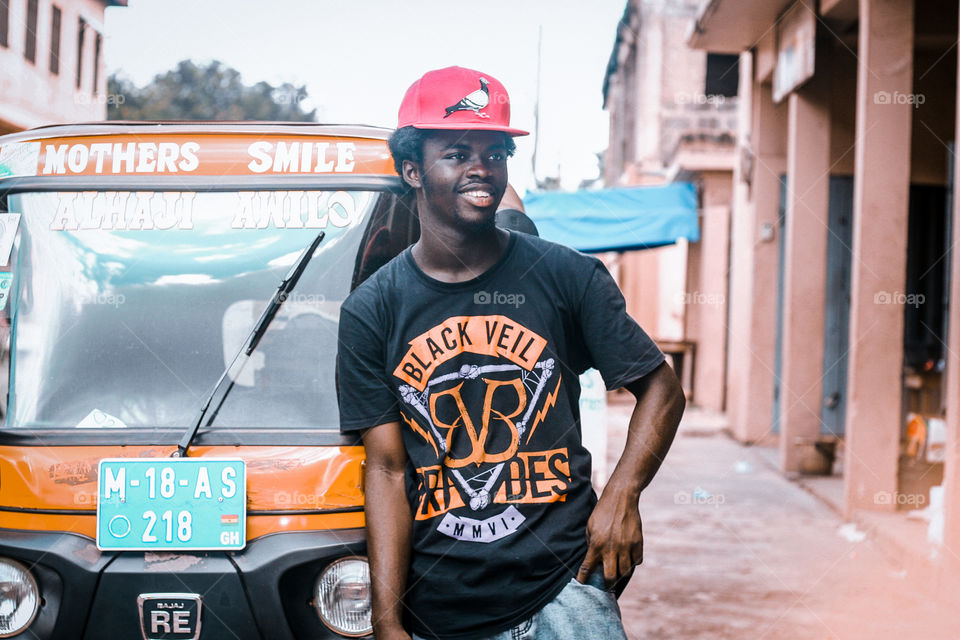 Young handsome looking teenager smiling standing in front of a yellow car with a red cap on his Afro head