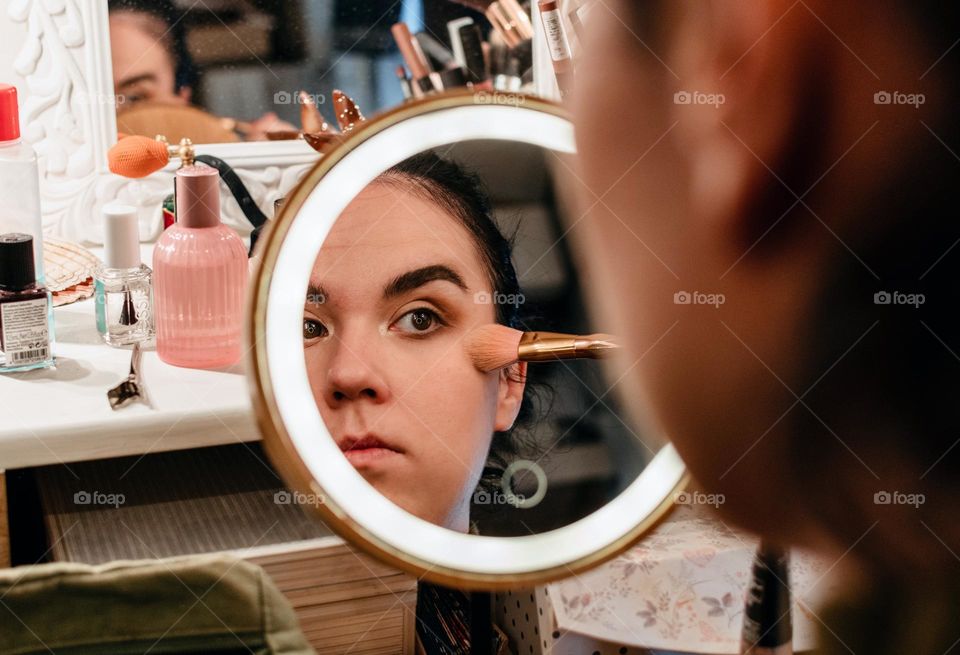 Mirror portrait of woman using make-up brush to apply make-up to her cheeks