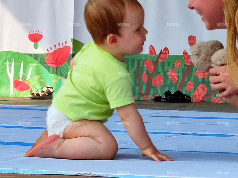Love: parents and children. A little boy in a diaper is sitting on the podium. The boy looks at his smiling mother, who is holding a teddy bear. The connection between baby and mother