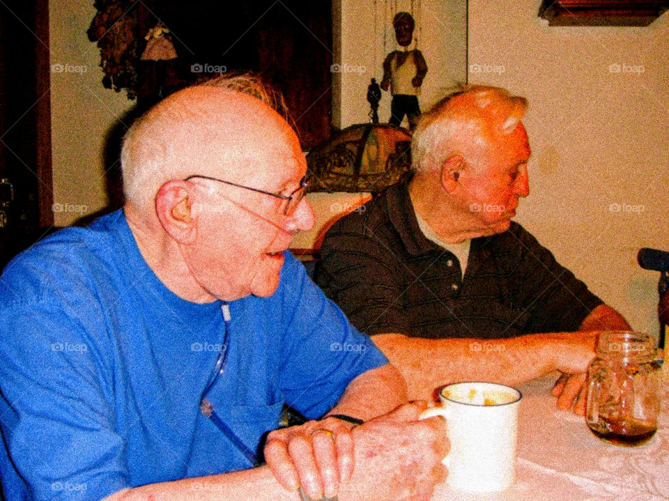Two elderly men sitting together at kitchen table, cup of coffee in hand & one grandfather needs to use oxygen. The men are balding with white hair & wears eyeglasses. They're loved family members & relatives.