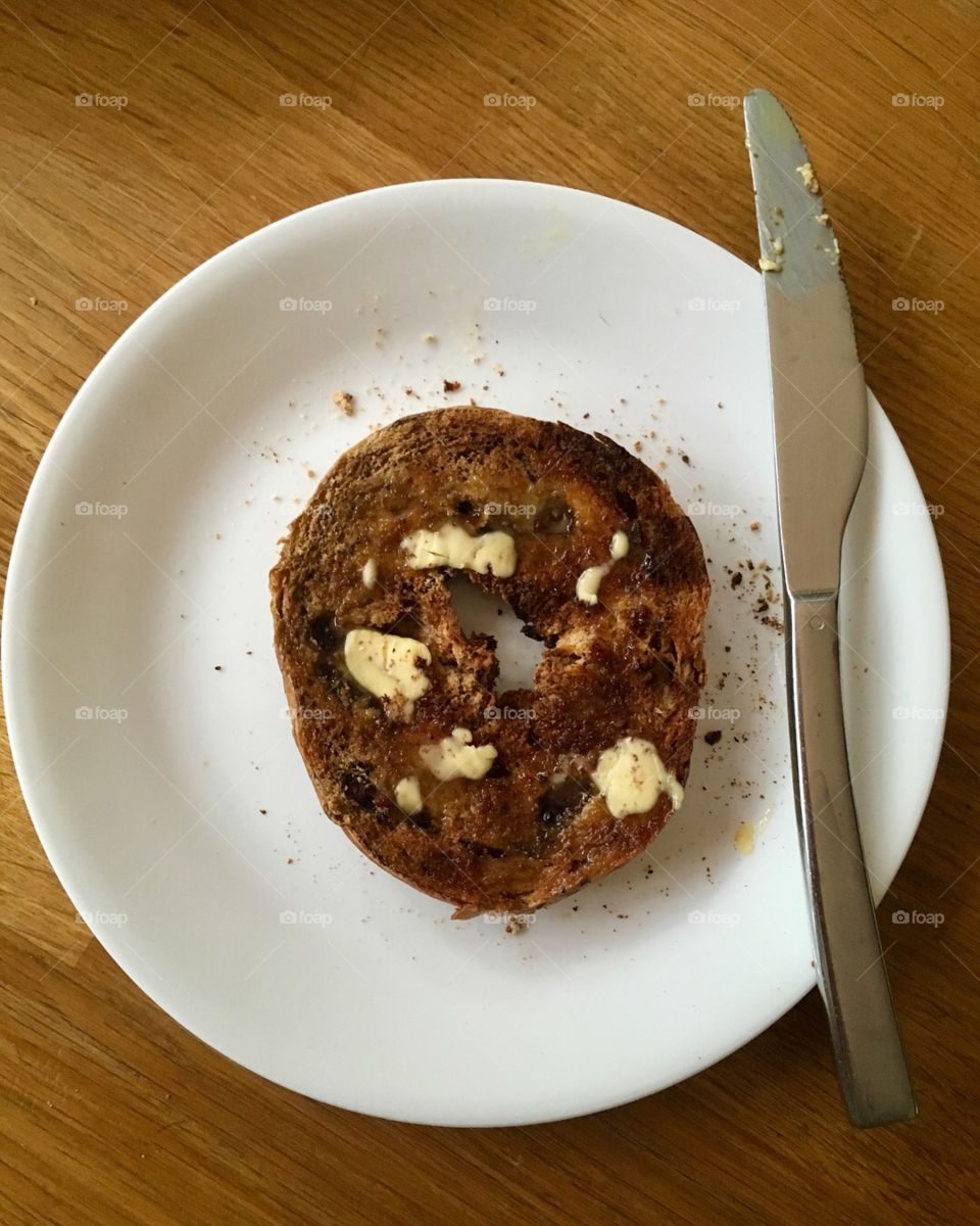 This is a photo of a toasted and buttered raisin bagel on a white plate on a wooden table.