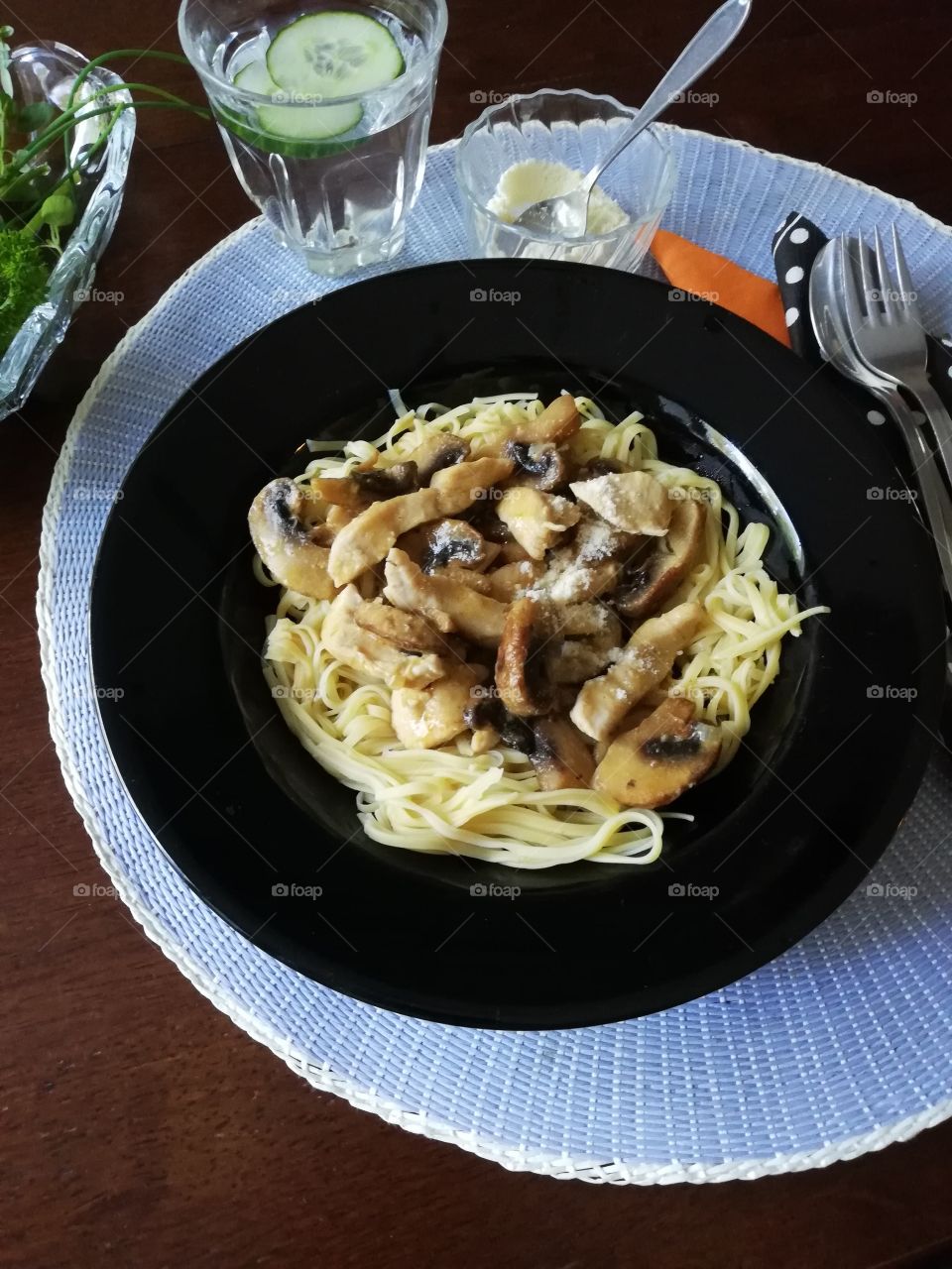 A black plate full of spagetti with chicken, mushrooms and parmesan on a light blue place mat. A glass of water with slices of cucumber and napkins, a fork, a spoon and a bowl of cheese on the table.