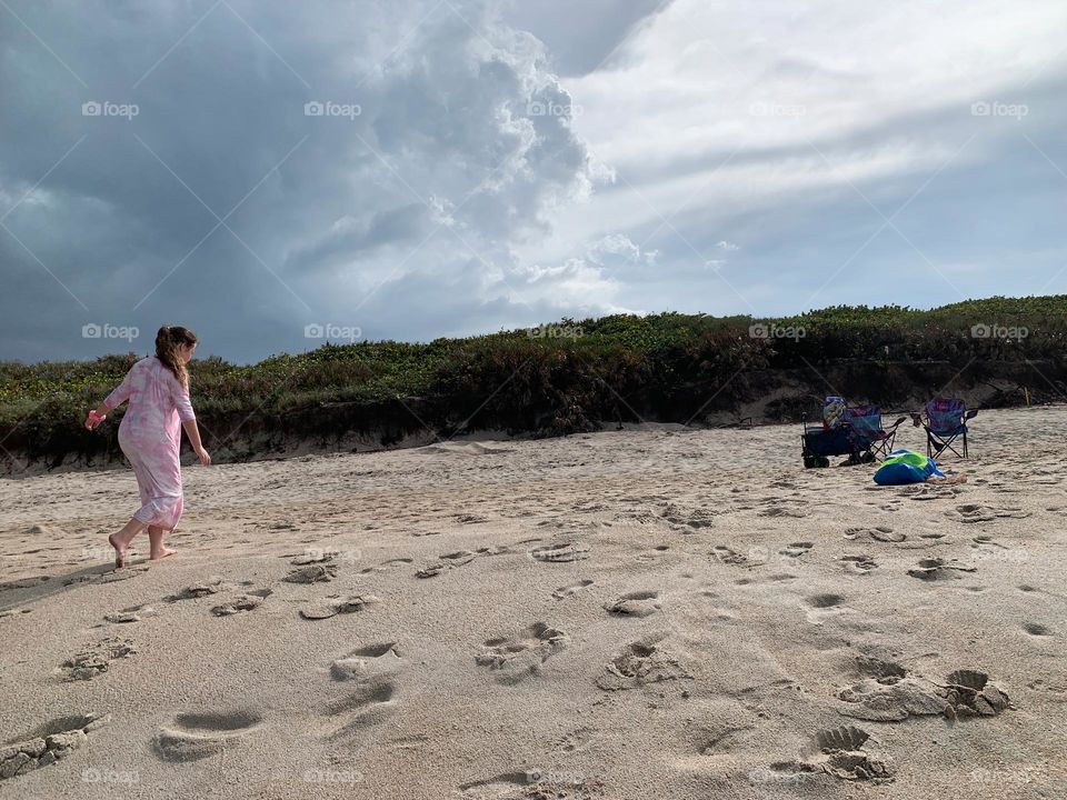 Child girl walking at the beach on the sand with multiple feet prints going towards the blue wagon and beach toys in a net bag with hill of tropical plants and grey cloudy sky on the background.