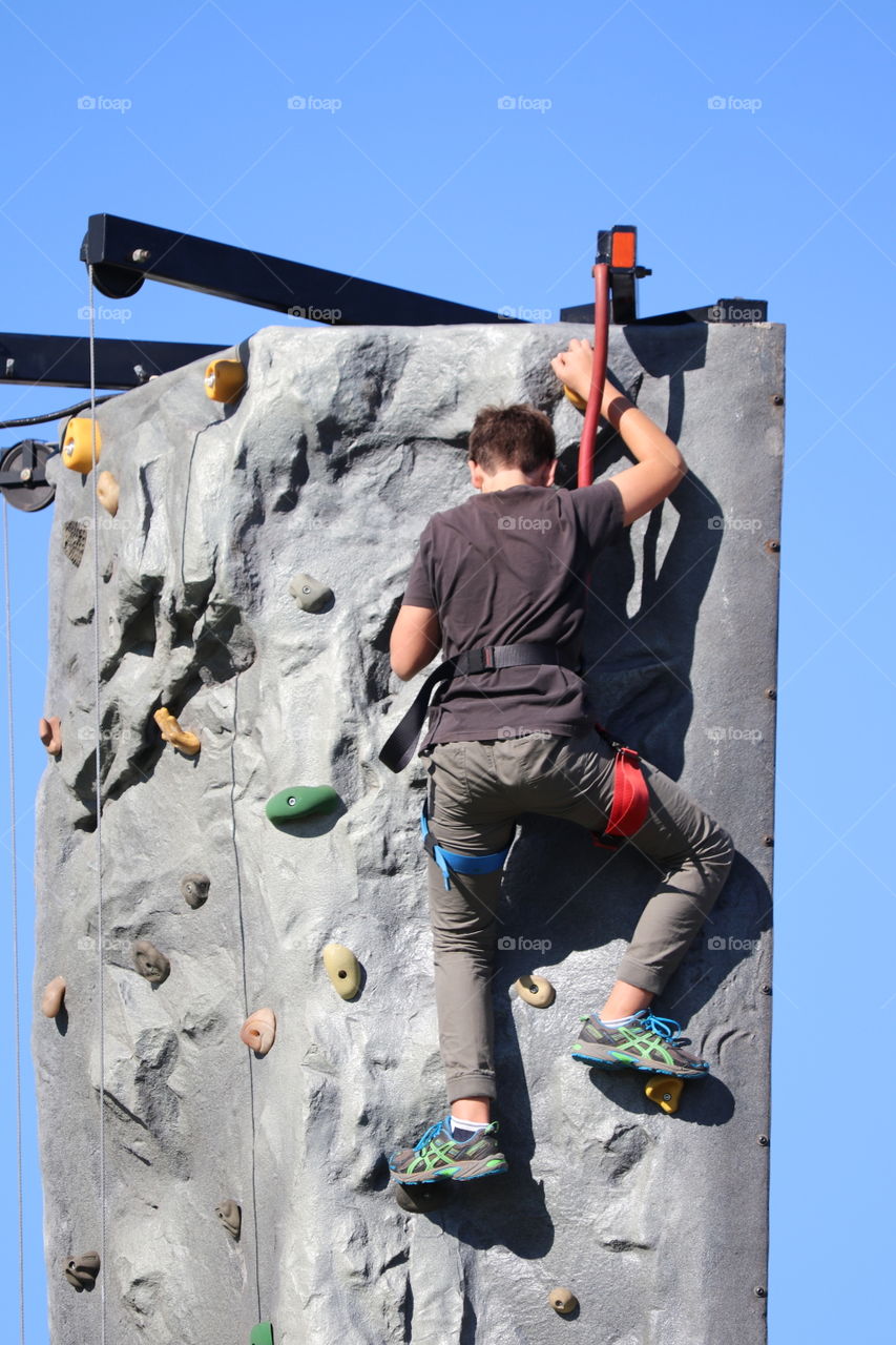 Young rock climber at top of rock climbing tower outdoors against a vivid blue sky. 