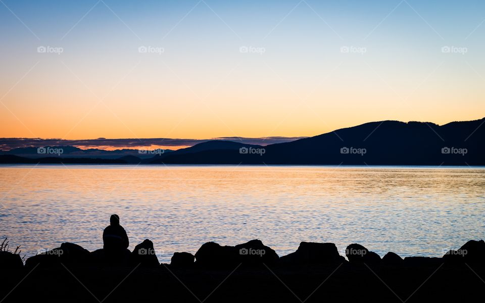 Watching the sunset. Silhouette of a person sitting on rocks watching the sunset over the water.