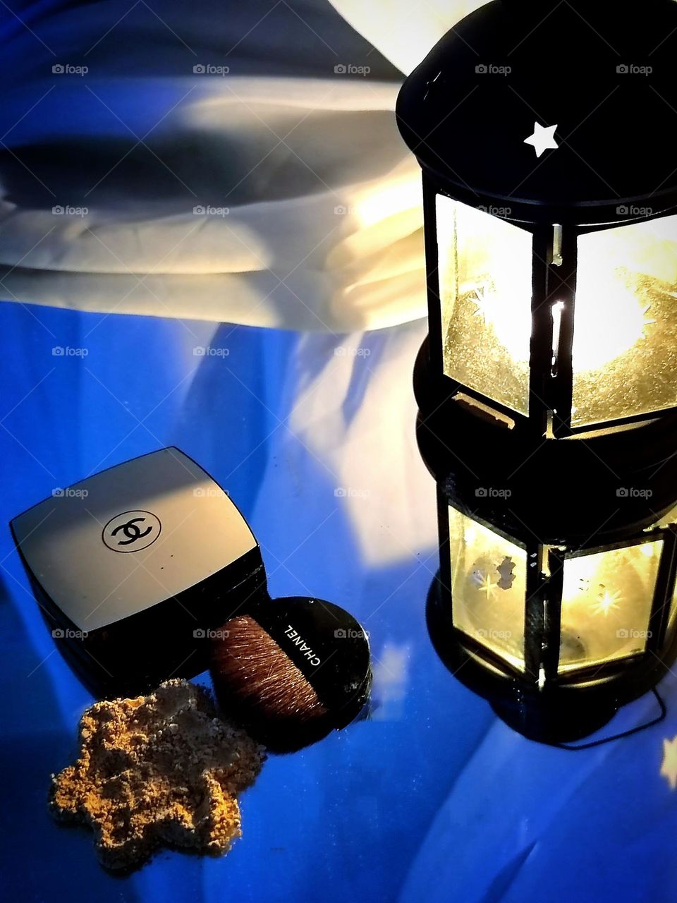 A lantern is lit against a mirror background, the light from which falls on a box with the Chanel logo, on powder and a brush with the inscription "CHANEL".  In the background is a white air curtain. Powder Chanel Les Beiges