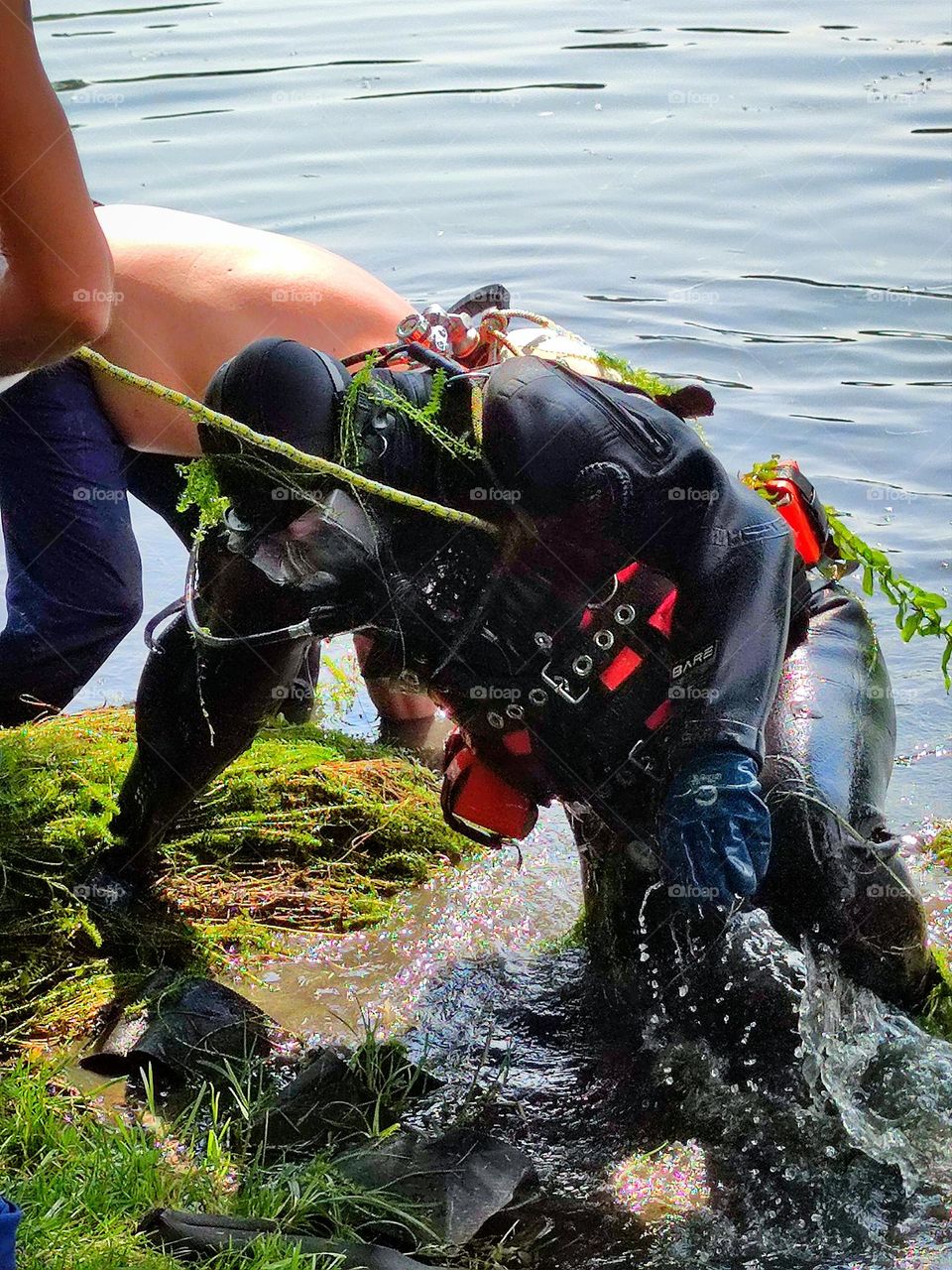 The scuba diver comes out of the river to the shore.  Green algae hang on a scuba diver.  On the shore they help him out of the water