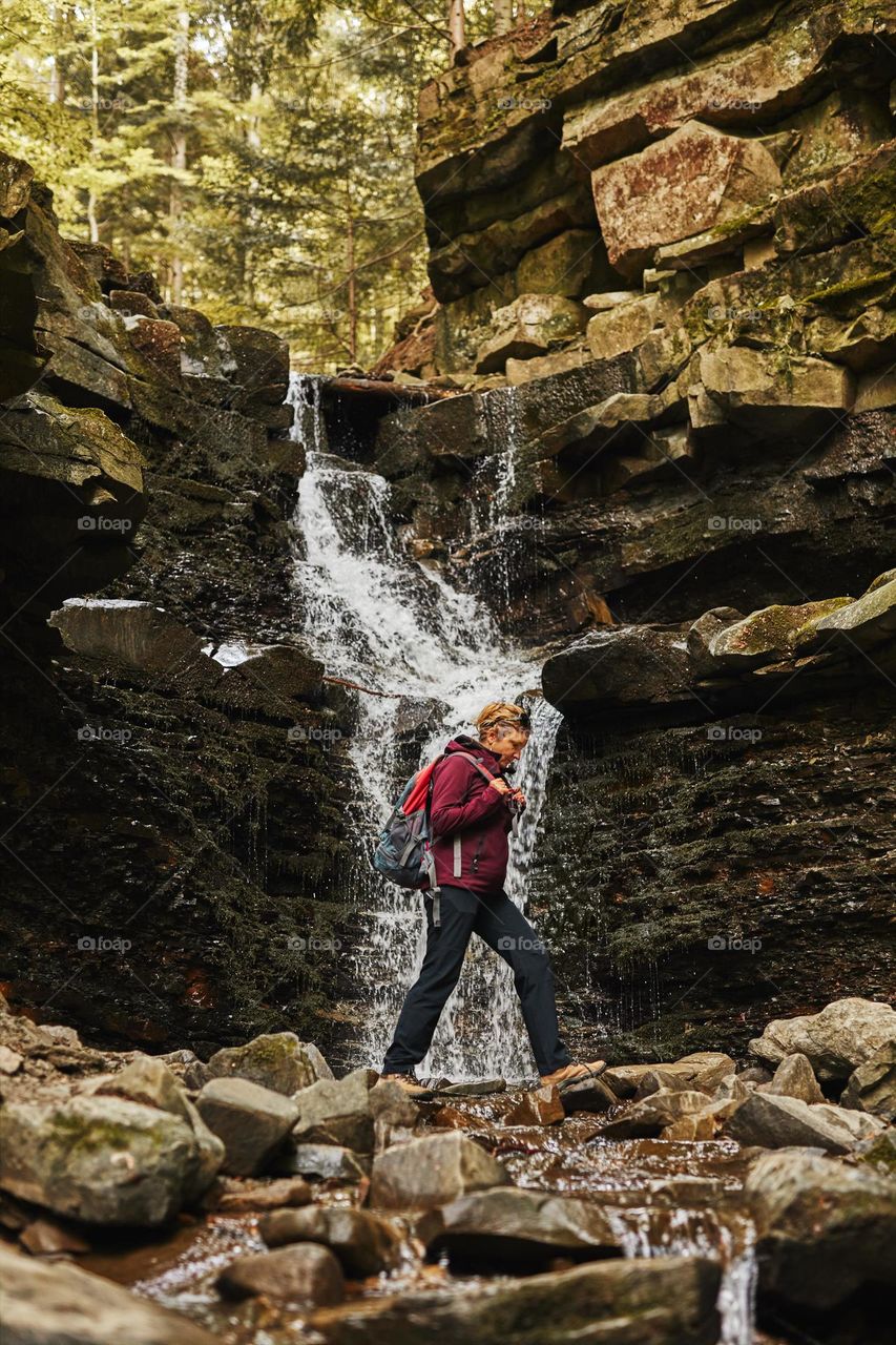 Hiking in mountains. Woman enjoying hike on sunny vacation day. Female with backpack walking close to waterfall. Spending summer vacation close to nature