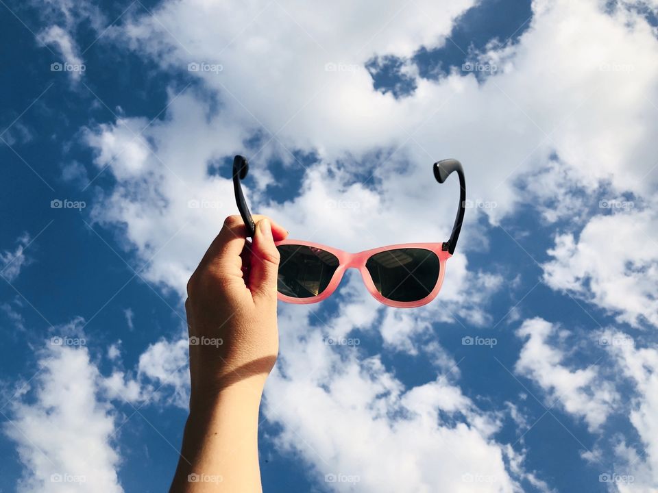 Hand holding pink sunglasses against summer blue sky covered with fluffy white clouds