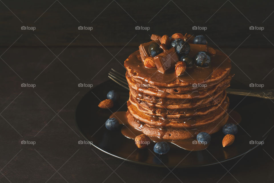 Stack of pancakes with chocolate sauce, blueberries and nuts on dark wooden desk