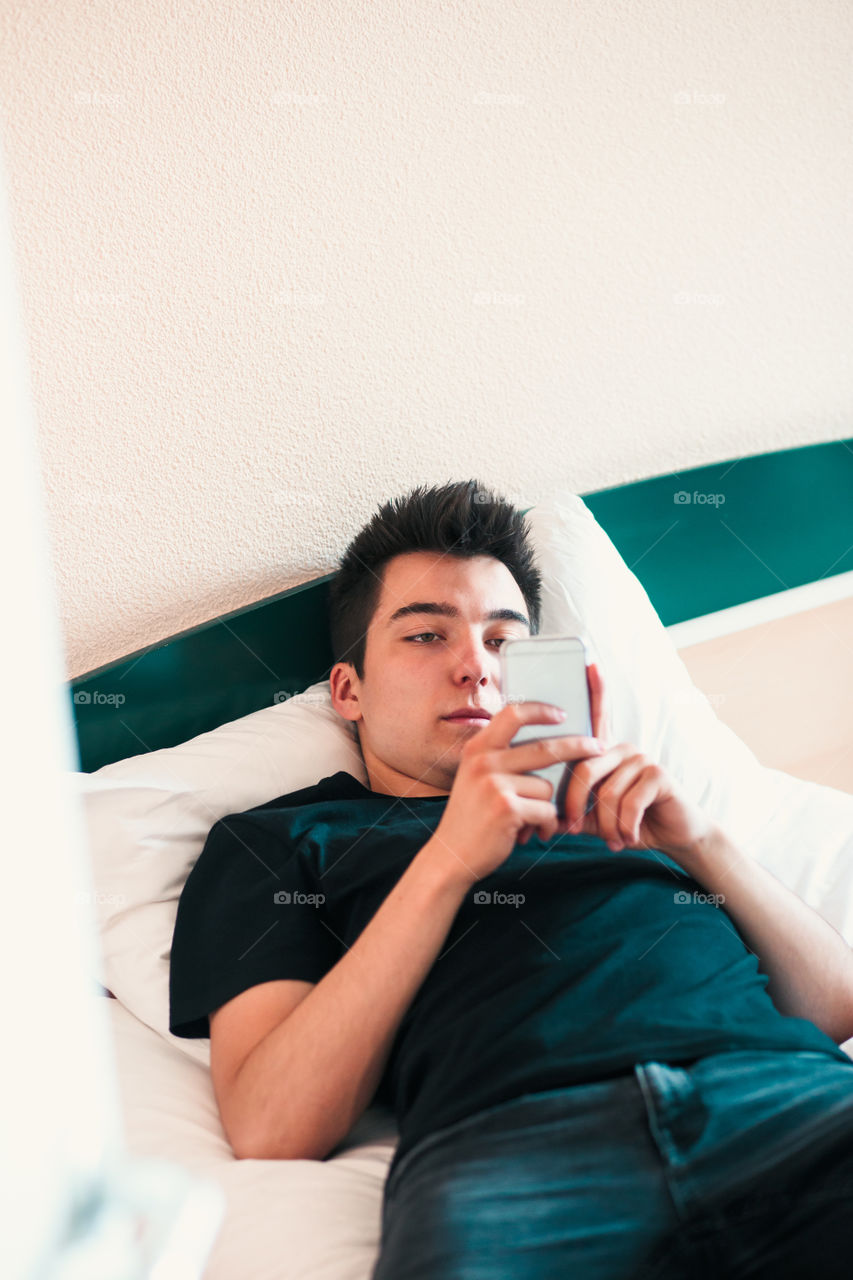 Young man using mobile phone laying in bed in bedroom