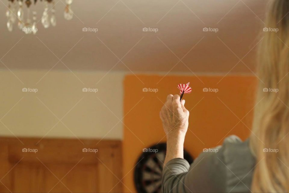 A woman from behind with long blonde hair holding a red dart and aiming at a dartboard in living room