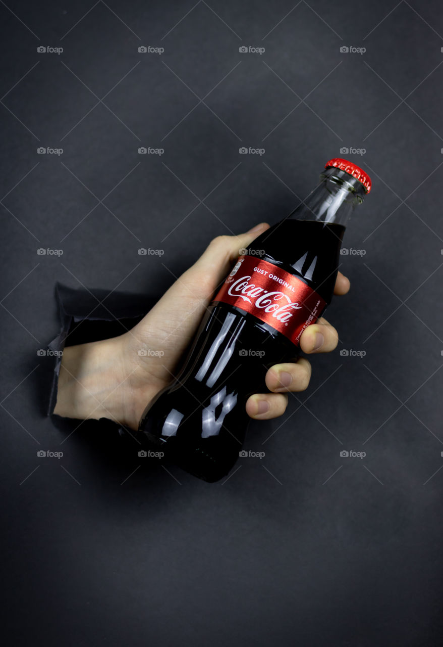 Hand with Coca-Cola bottle in paper hole on dark background.