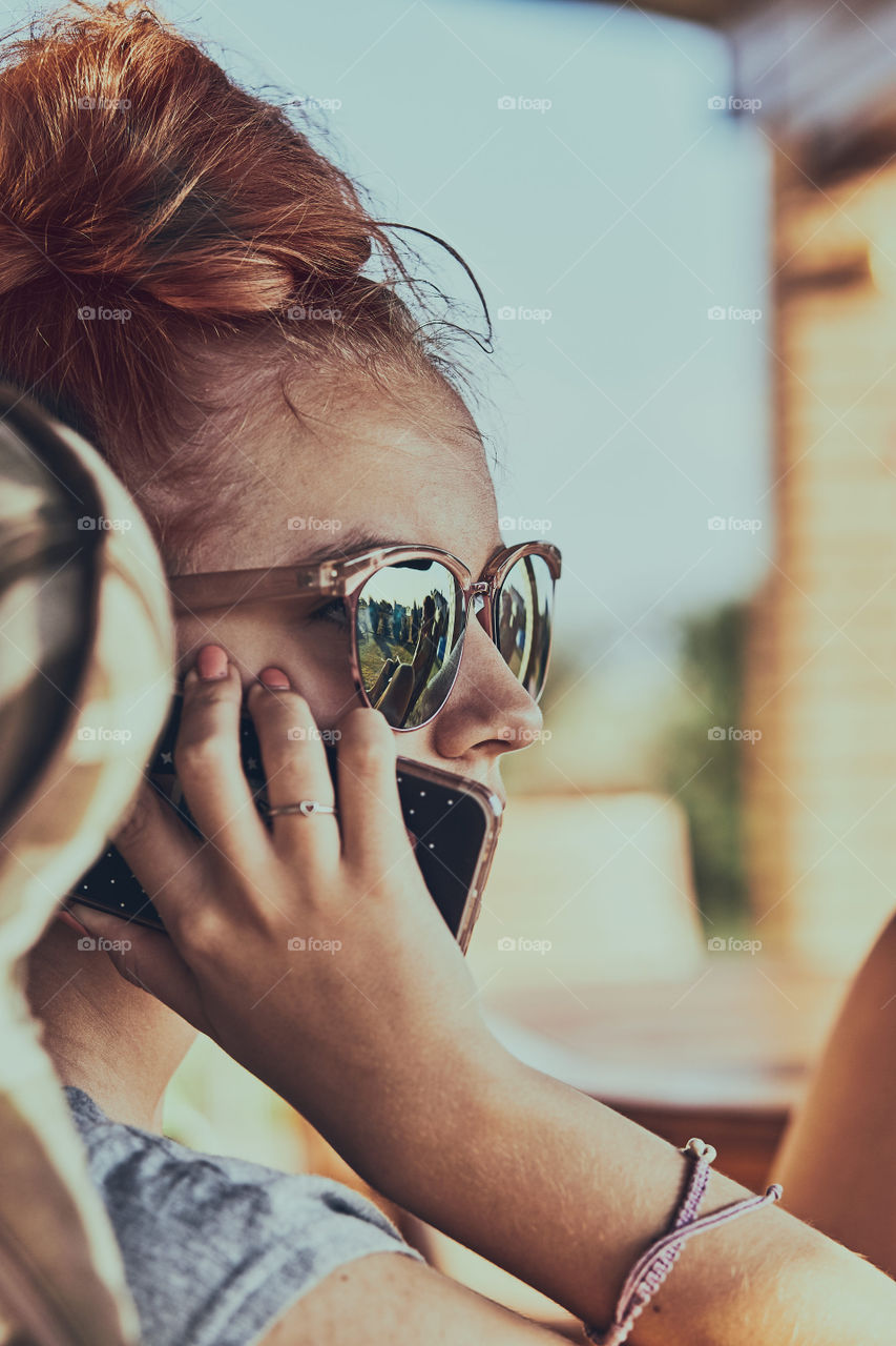 Young woman talking on a  mobile phone sitting in a chair outdoors on patio wearing a sunglasses. Real people, authentic situations