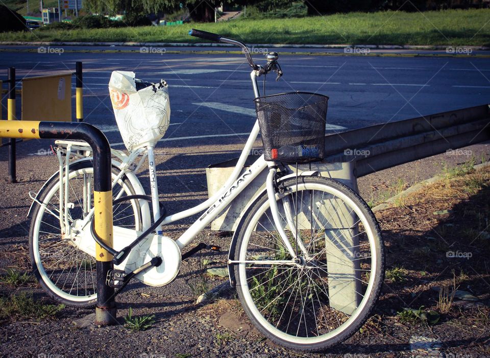 One white lonely bicycle with a bag over the seat and basket locked to a metal fence.