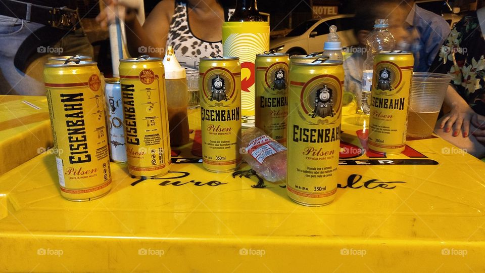 Latinhas de alumínio. Cerveja Eisenbah em mesas amarelas. Pessoas em confraternização.