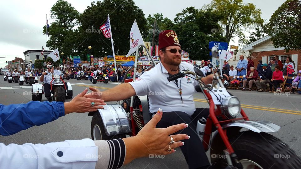 Shriner in a parade riding a trike entertains the crowd and slaps their hands as he drives by.