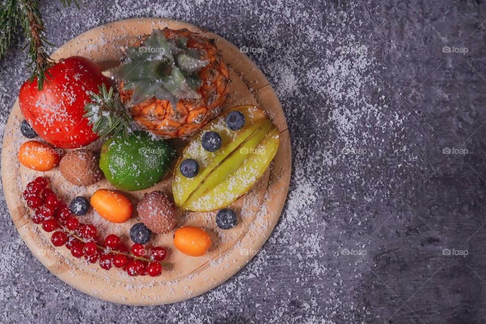 Tropical fruits and berries from pineapple, currant, lime, blueberry, mini tangerines, persimmon and lychee on a round wooden tarkle lie on the left on a dark cement background with copy space on the right, flat lay close-up. Tropical fruits and berr