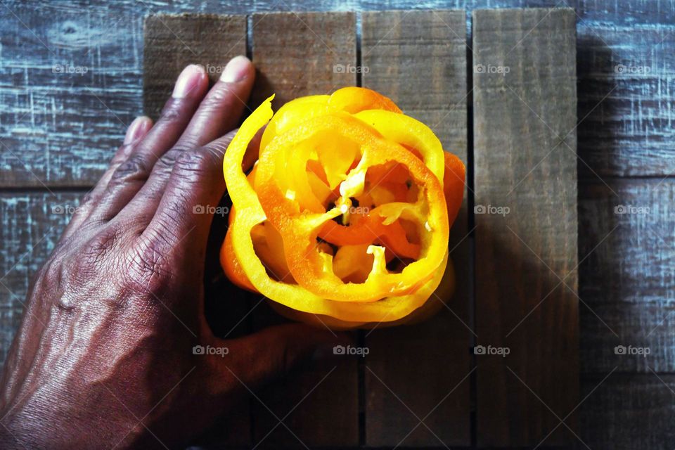 A man arranges slices of a yellow bell pepper in preparation for a meal from a flat lay point of view on a wooden cutting board.