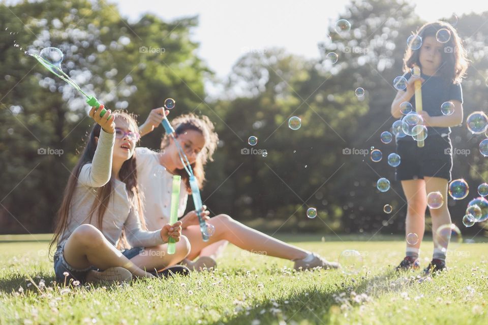 Three beautiful, happy Caucasian sister girls sit on a green lawn in a public park and blow bubbles with joyful real emotions, close-up side view. The concept of parks and playgrounds, children's entertainment, outdoor recreation, children's picnic,