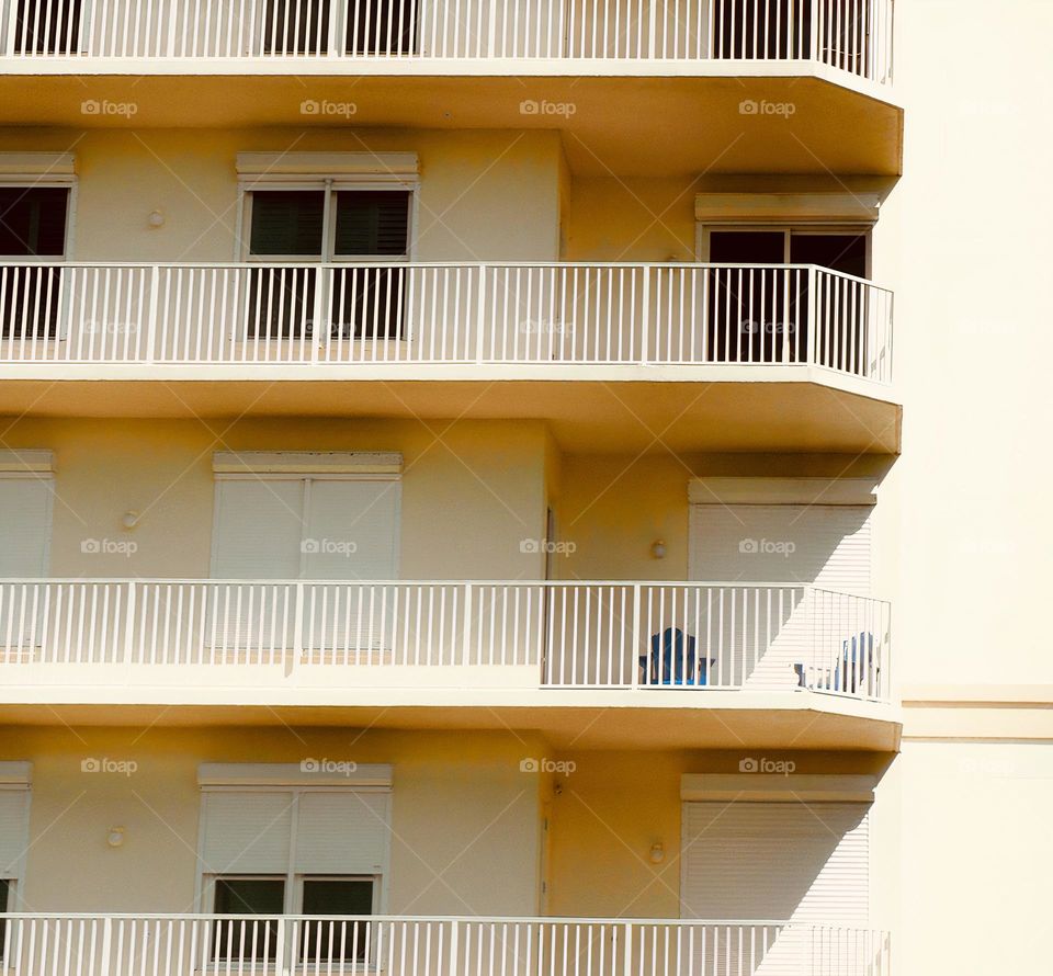 Symmetrical apartment’s units creating a geometrical effect with light and shadows with fenced patios with banisters, handrails, banisters, looking at windows and blinds or curtains, doors and chairs.