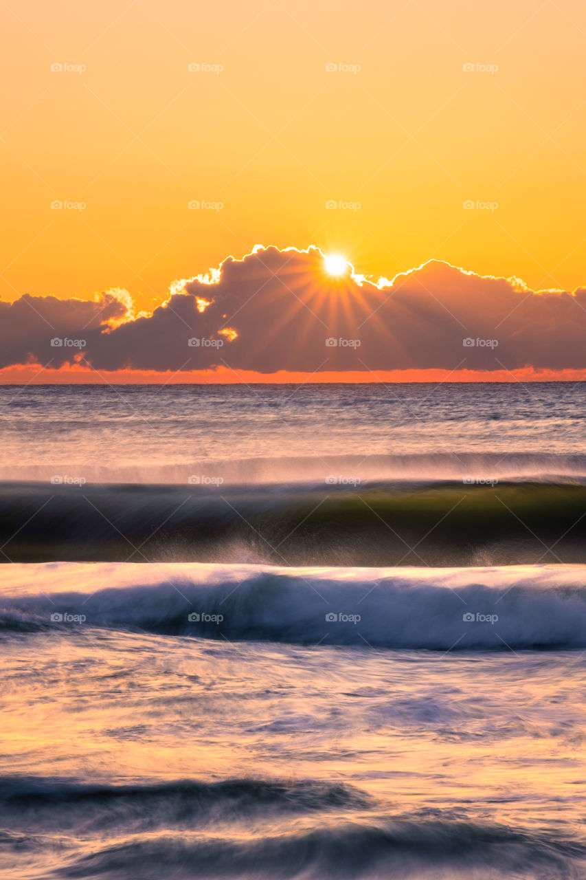 The rising sun popping out from being a cloud creating a star burst, with golden light illuminating waves in the ocean 