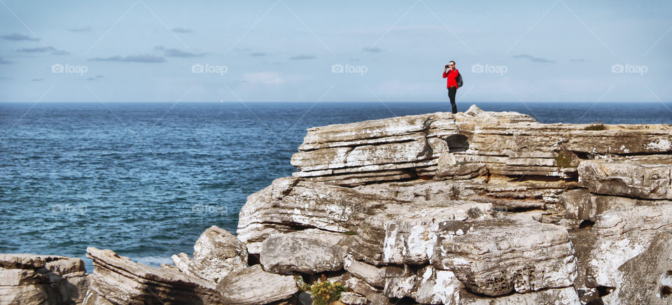 A man stands out in his red shirt as a solitary figure standing on a cliff edge, as he takes pictures of the coastline 