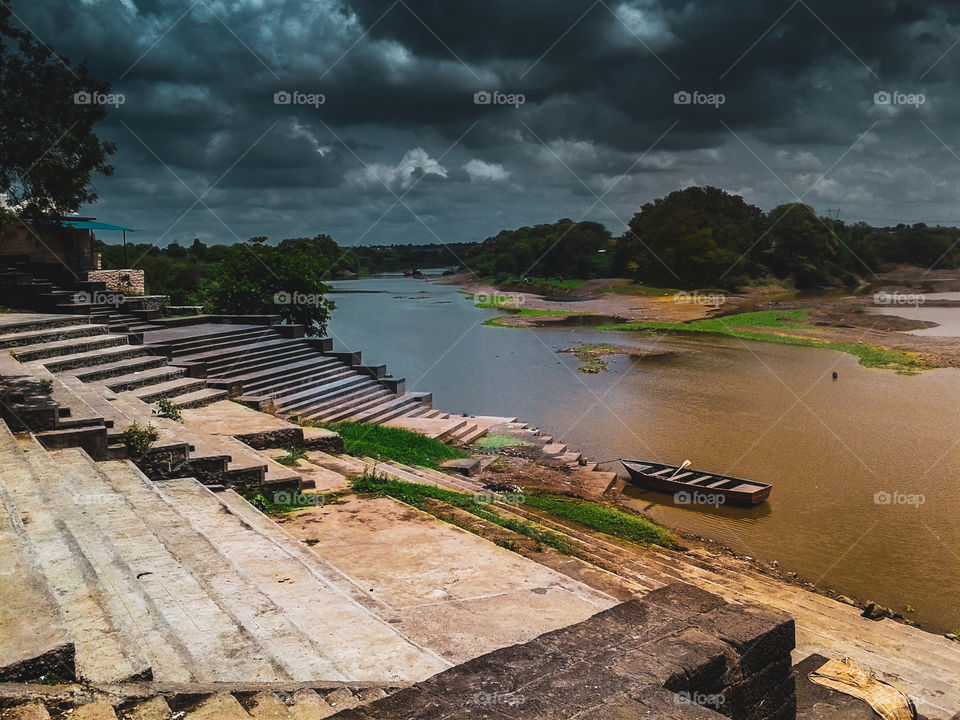 Landscape - it is bank of river with stairs and boat in water. There is a cloudy sky with reflection in river.