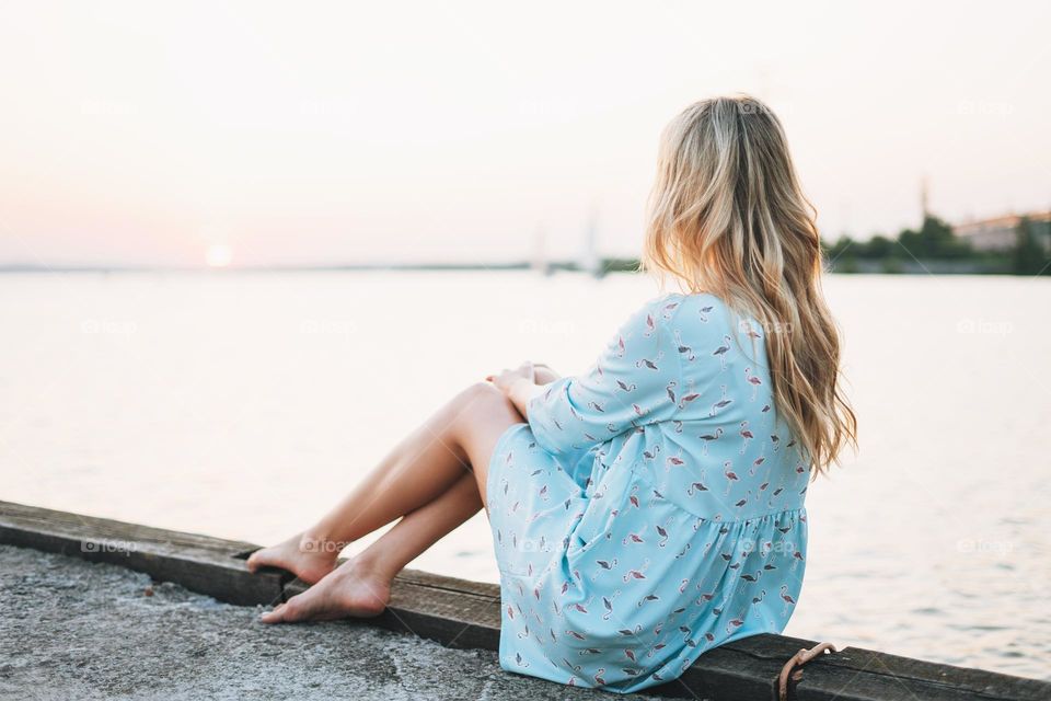 Beautiful blonde young woman in blue dress sitting on pier and looking on sunset