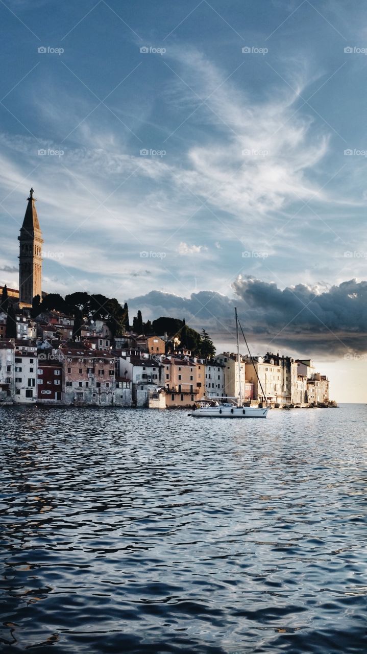 The buildings of Rovinj, Croatia - including the tower of the Church of St Euphemia - lit by the falling sun at sunset with a boat in the sea.