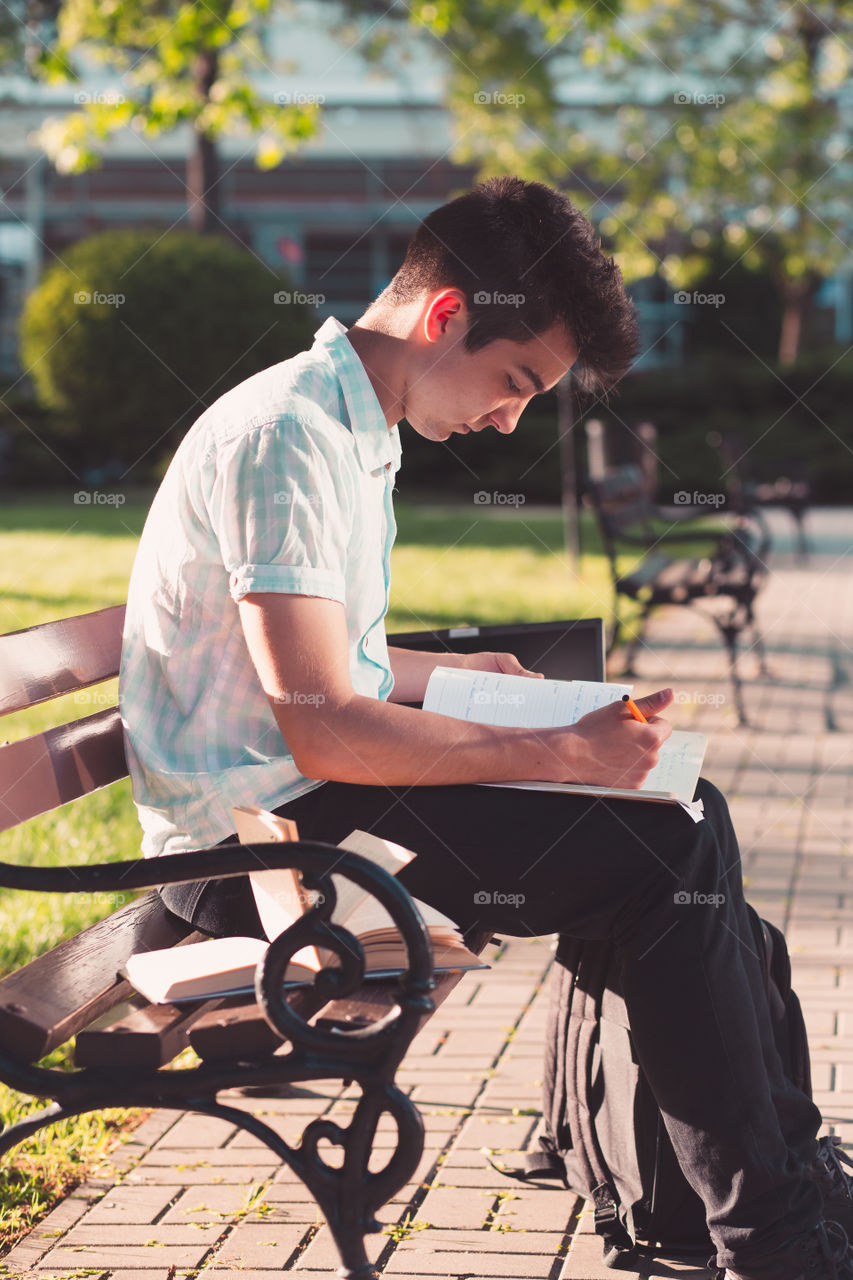 Student working on a laptop using books and notes sitting on a bench in a park. Young boy wearing a blue shirt and dark jeans