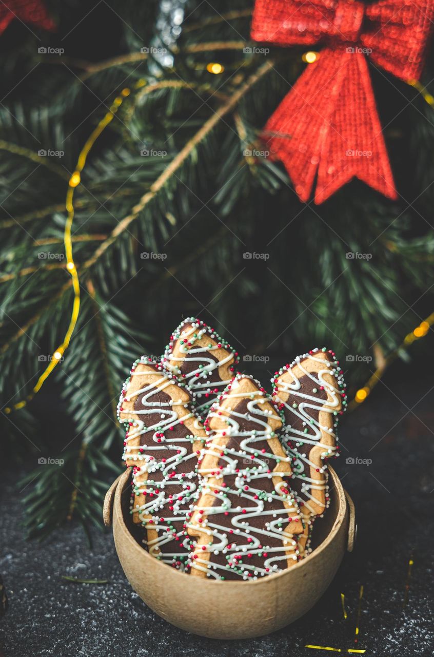 Shortbread cookies in the shape of Christmas trees in a craft natural bowl stand on a dark background with spruce branches and 7 cornflowers, close-up side view.