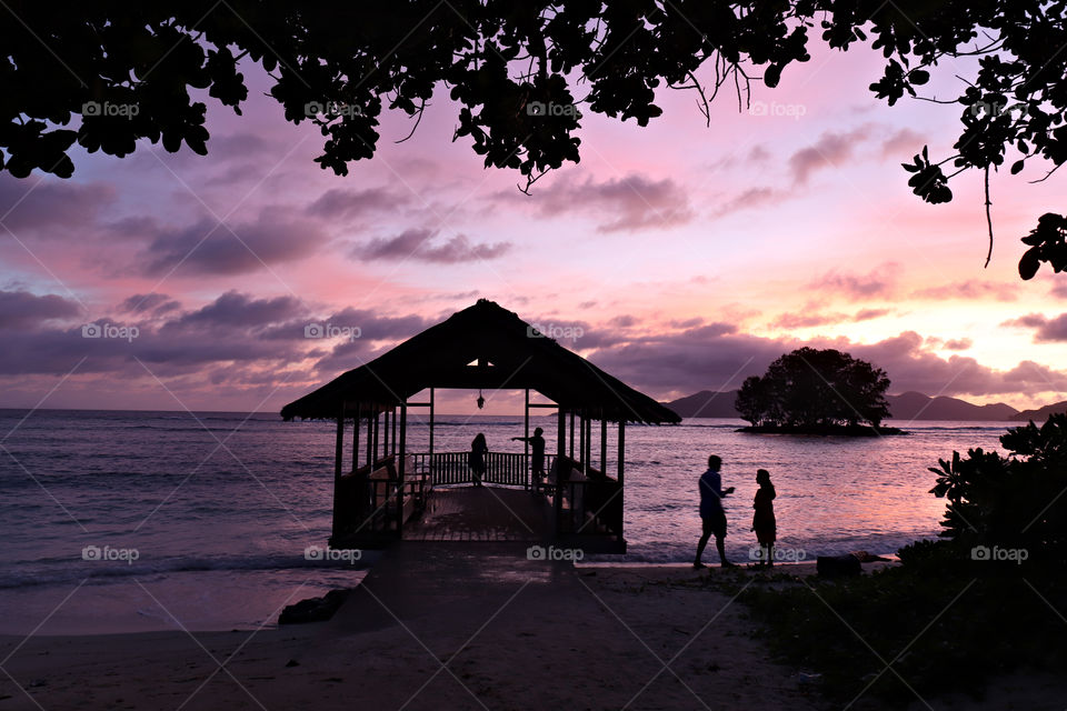 Silhouetted Family enjoying sunset at little beach hut next to the ocean framed by leaves.