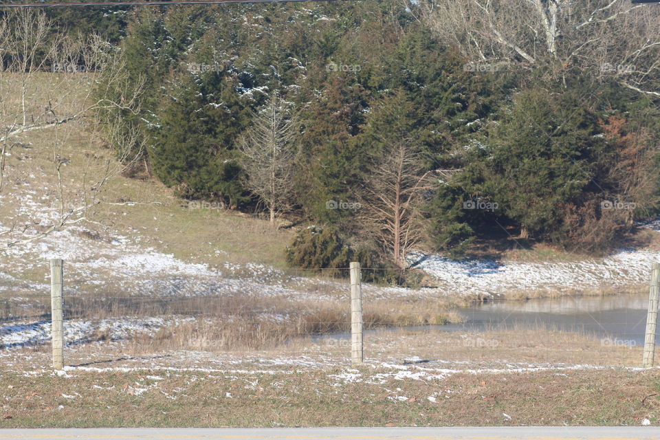 A wintertime view of the land, including a partially frozen pond. A fence is visible in the foreground. The sun is shining from behind the photographer.
