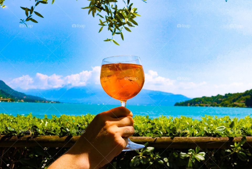 Hand holding aperitif spritz, typical Italian drink, facing the lake, with beautiful landscape in the background, blue sky and sunny