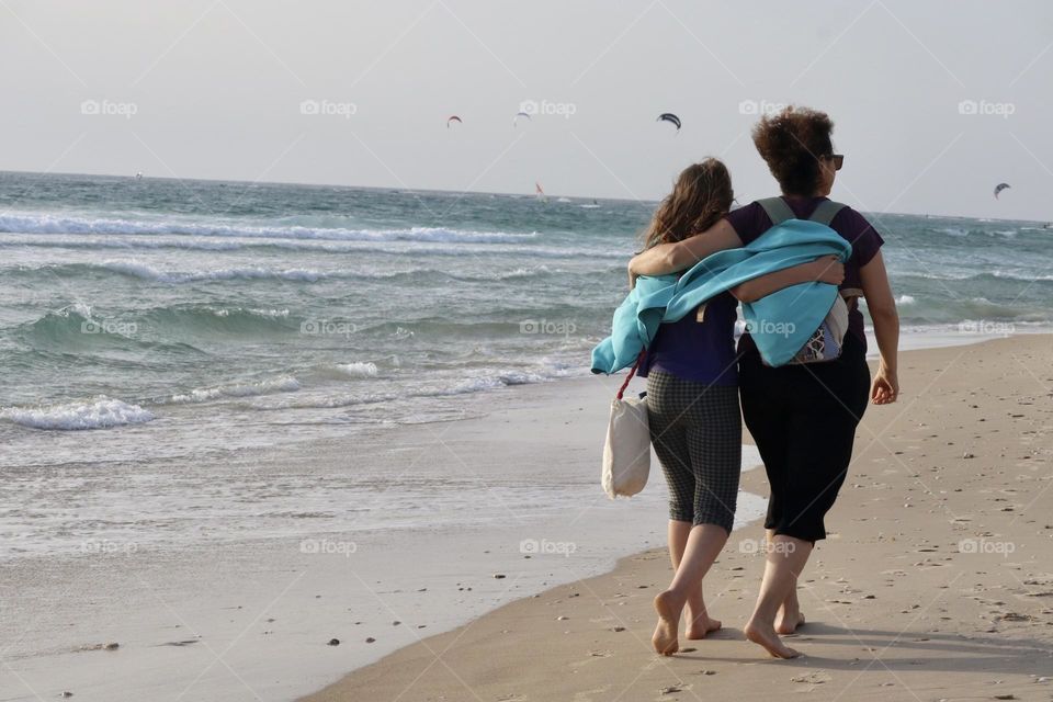 Mother and daughter walking along the beach hugging 