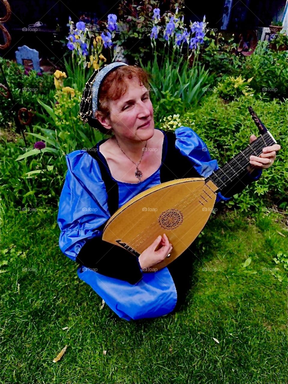 This lovely lady has a flair for bygone ages. She wears a Renaissance to medieval inspired theatrical gown and plays a custom built lute after a historical model in this beautiful garden in “Lower Bavaria”, Germany. 2024. Hypnotic Productions