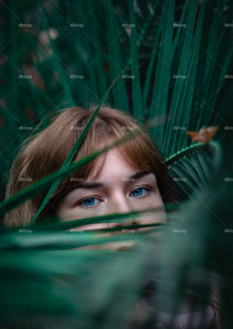 Blue eyed and blonde girl peeking out of the palm leaves.