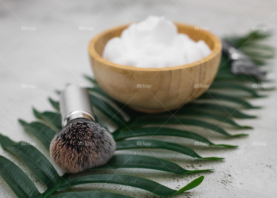 Fluffy shaving brush made of natural fleece, wooden bowl with shaving foam and razor lie on a green palm branch on a light stone blurred background,side view. The concept of barbershop men, shaving men,beard care.