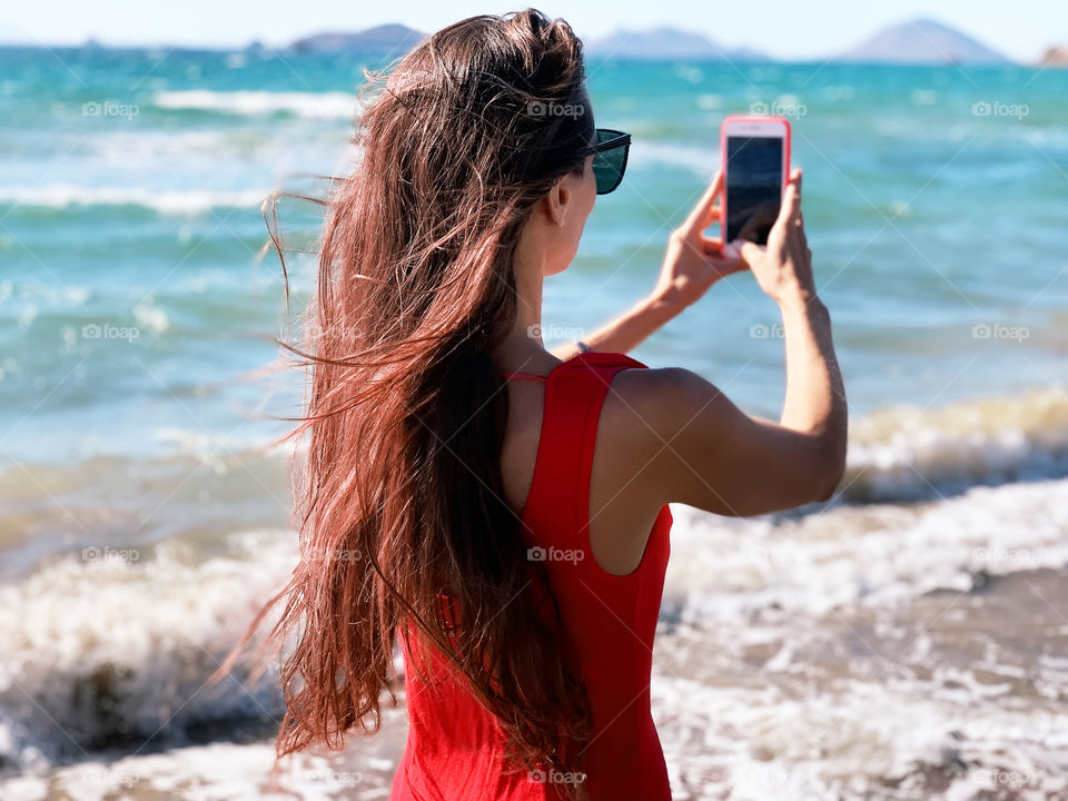 Young woman with long red hair in red dress taking a photo using red mobile phone at the seaside 
