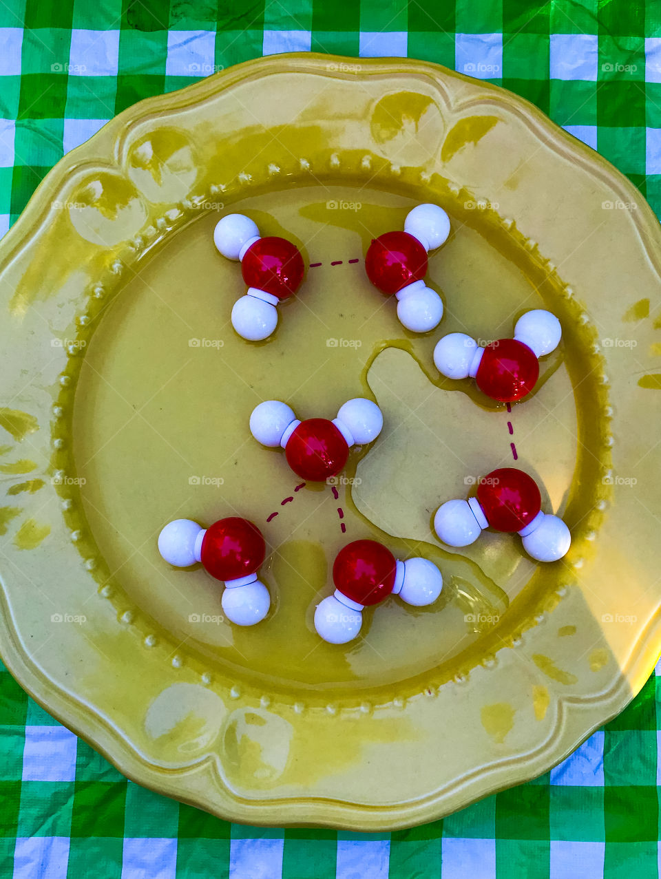 Clash of Colours: Chemistry geek alert! Red (Hydrogen) & white (Oxygen) molecule model of two or three molecules representing liquid H2O(water), in a pool of water, on a yellow plate, on a green plaid tablecloth draped table. 💧