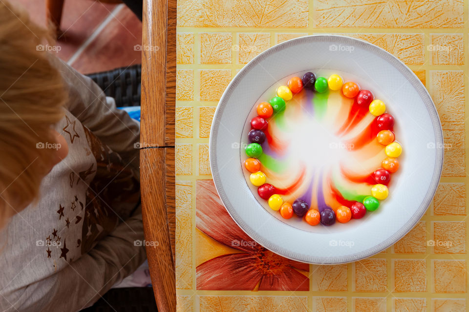 Child experimenting with sweet Skittles fruit pastilles creating rainbow on a plate by adding a drop of water.